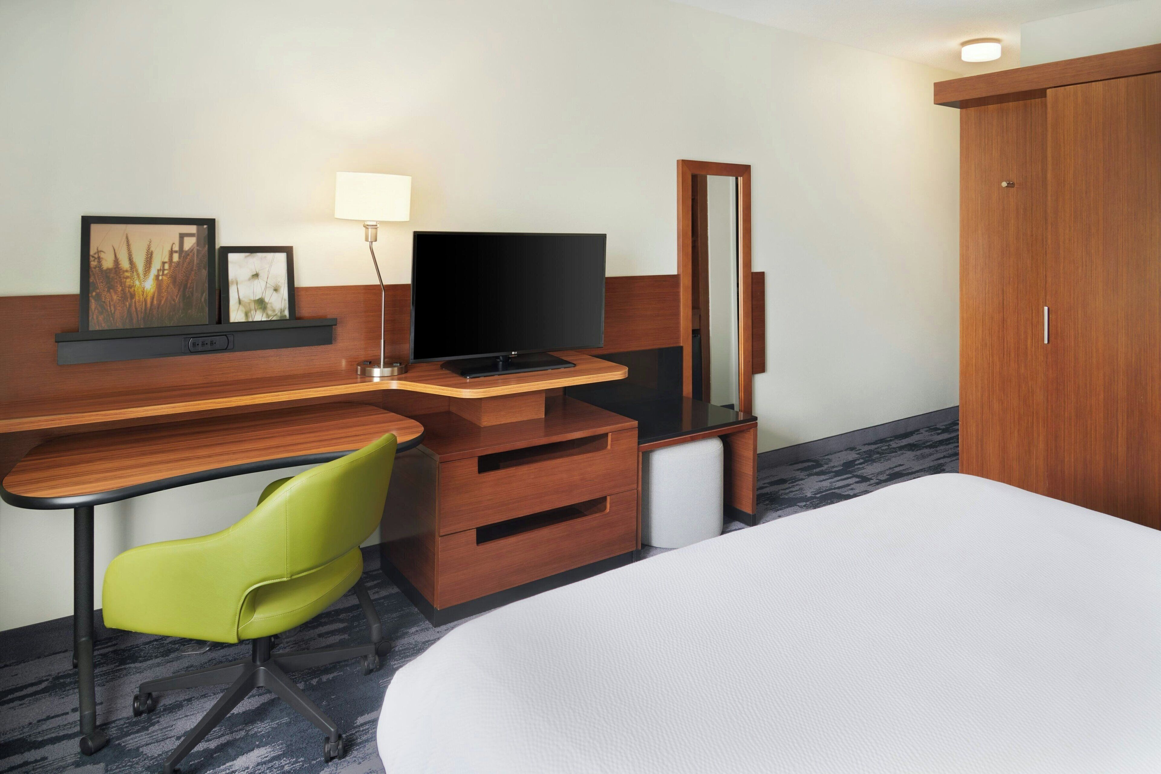 A hotel room with a wooden desk, a television, a green chair, a bed, and a wardrobe.