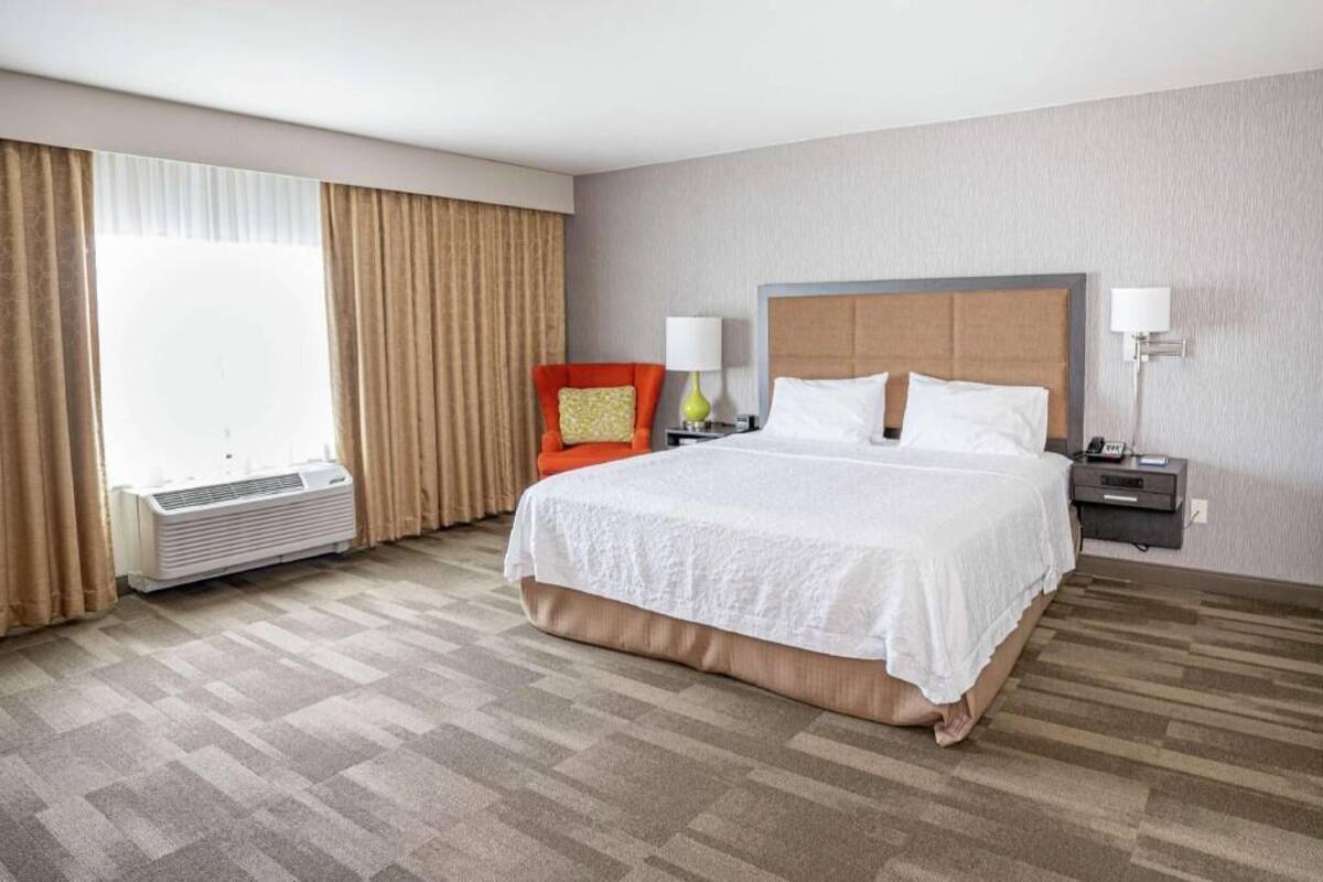Quarto de hotel com uma cama grande, ar-condicionado, uma cadeira e uma mesa de cabeceira com abajur.