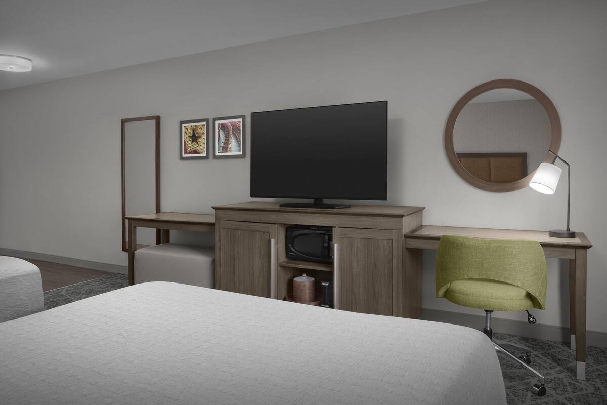 Quarto de hotel com uma cama, uma escrivaninha com televisão, uma cadeira, um espelho e quadros nas paredes.