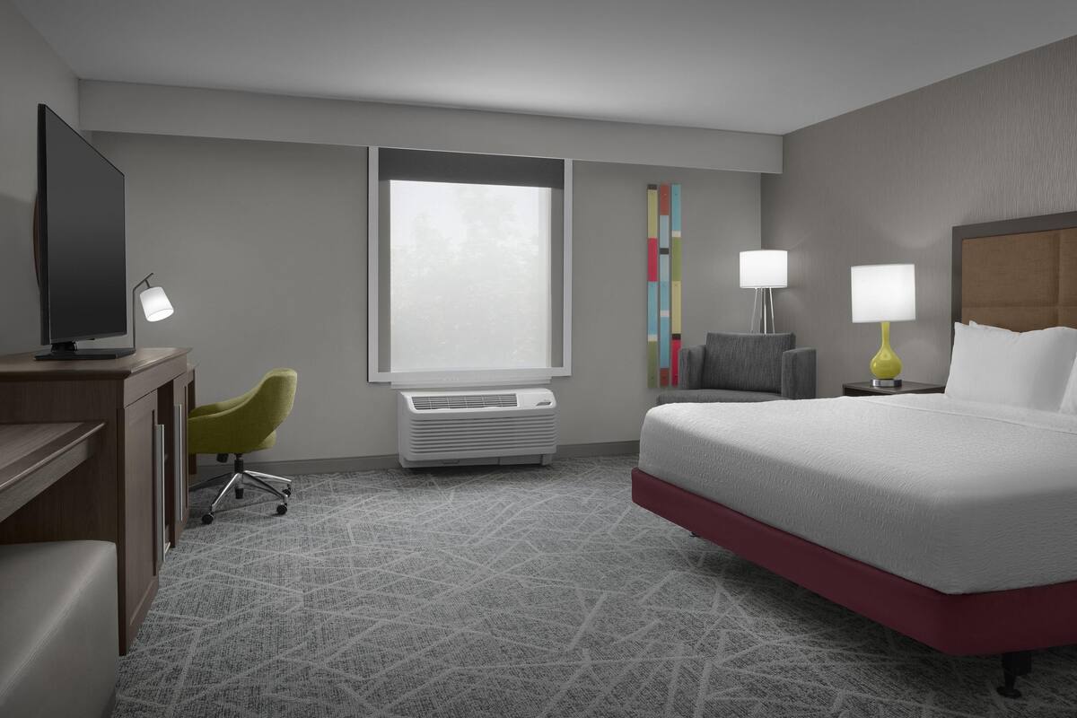 Quarto de hotel com cama, escrivaninha com televisão, cadeira, duas luminárias de cabeceira, janela e ar condicionado.