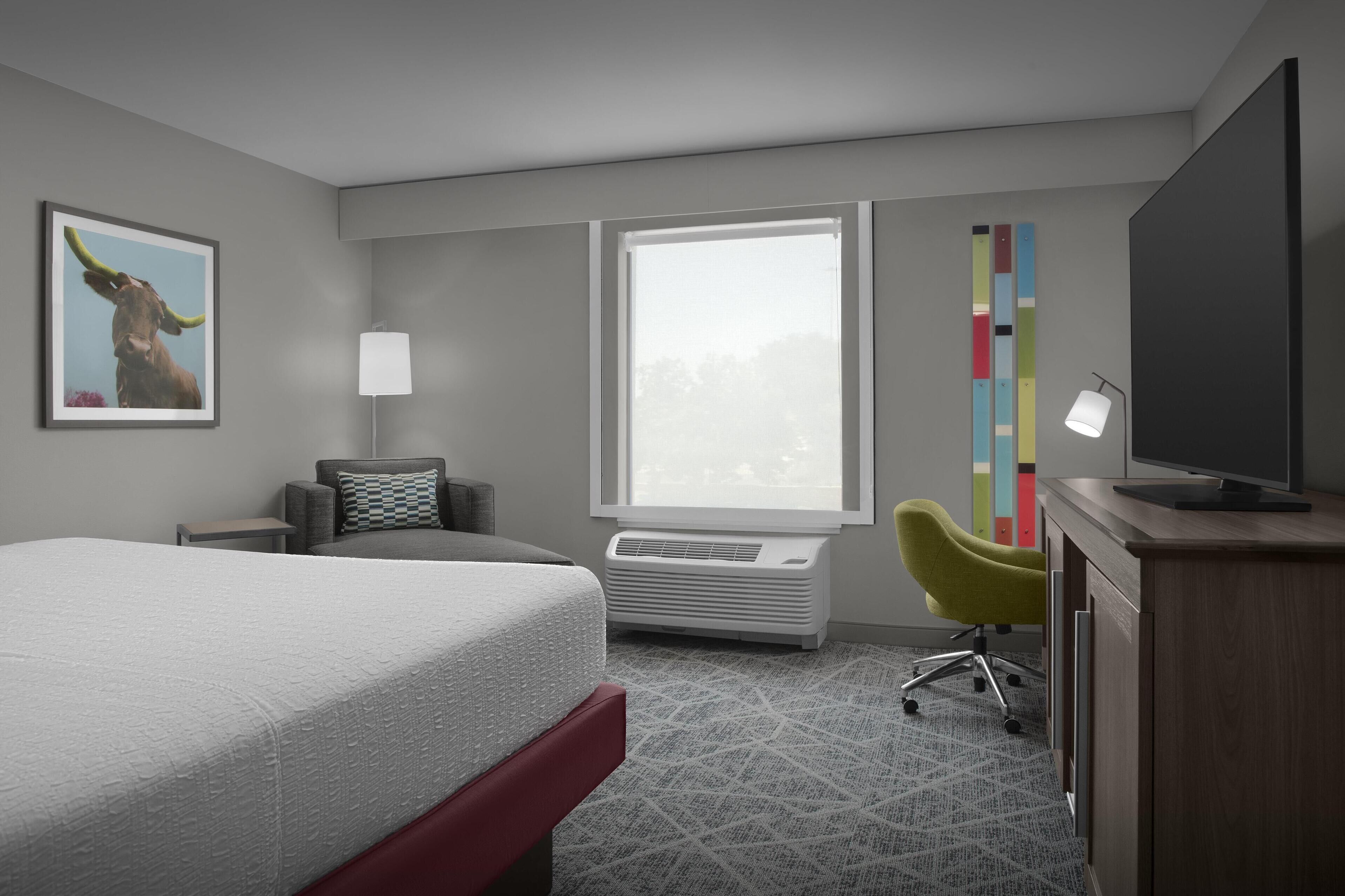 A hotel room with a bed, a desk, a chair, a TV, a window, and a colorful wall decoration.