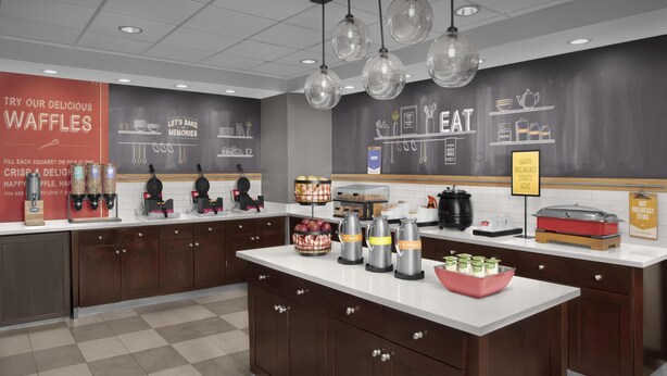 A modern kitchen with waffle stations, coffee makers, and a chalkboard wall with cooking illustrations. - Hampton Inn & Suites Georgetown/Austin North (Georgetown)