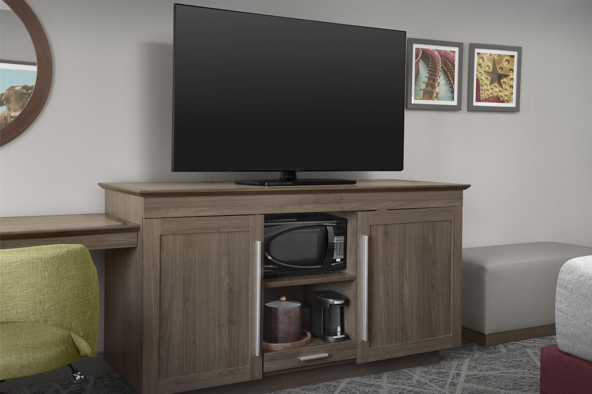 Quarto de hotel moderno com uma televisão de tela plana fixada acima de um armário de madeira. O armário contém um micro-ondas e uma cafeteira. Há uma cadeira verde e um banquinho vermelho no quarto.