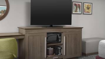 A modern hotel room with a flat-screen TV mounted above a wooden cabinet. The cabinet contains a microwave and a coffee maker. There is a green chair and a red ottoman in the room.