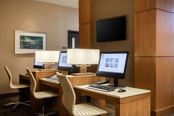 Ein Hotelzimmer mit drei Schreibtischen, jeweils ausgestattet mit einem Computer, Stuhl und einer Schreibtischlampe. Ein Flachbildfernseher ist über den Schreibtischen an der Wand befestigt.