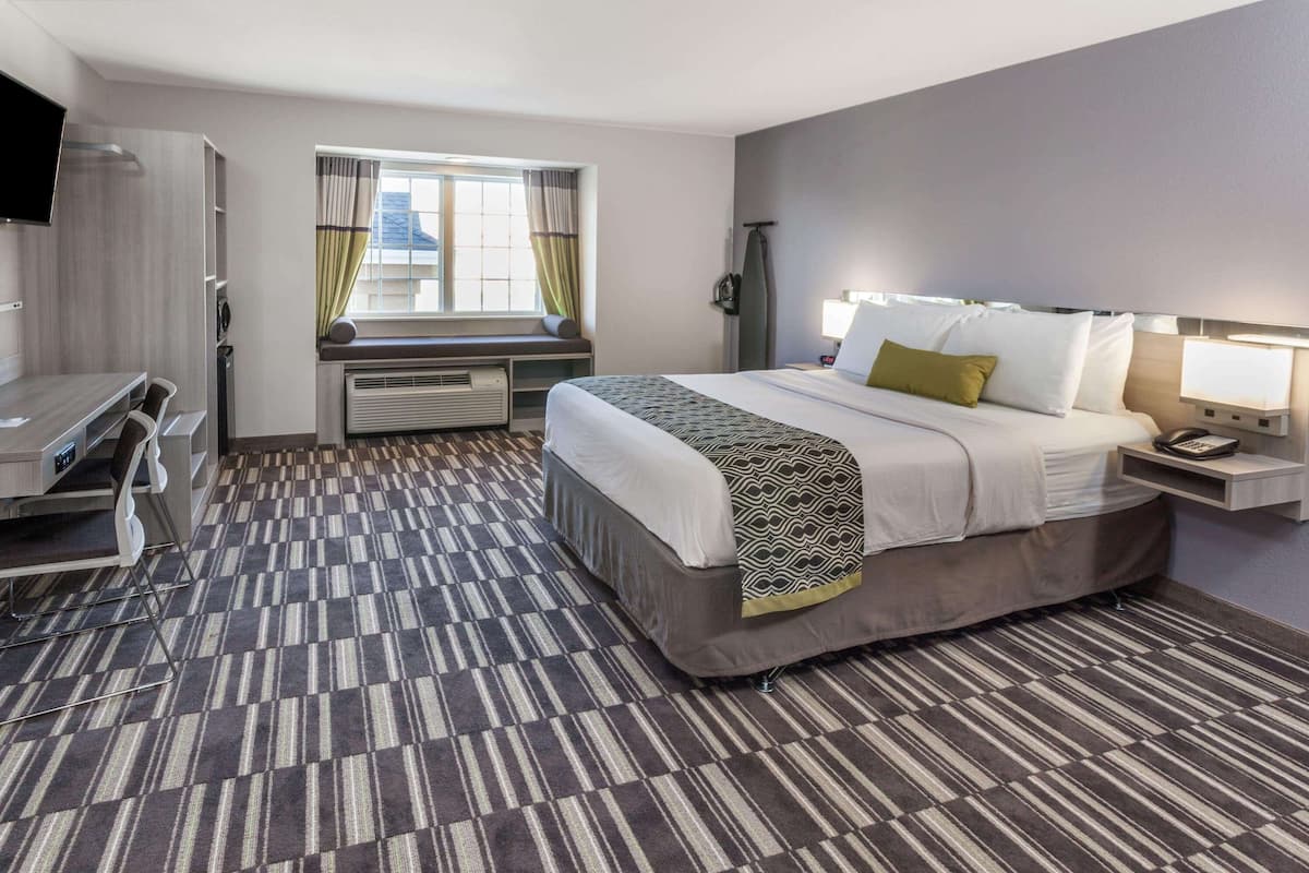 A hotel room with a bed, a desk, a chair, a TV, a window with curtains, and a wall-mounted lamp.