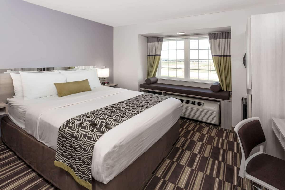 A hotel room with a large bed, a desk, a chair, a window with curtains, and an air conditioning unit.