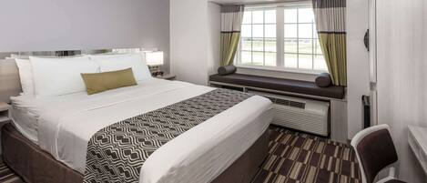 A hotel room with a large bed, a desk, a chair, a window with curtains, and an air conditioning unit.