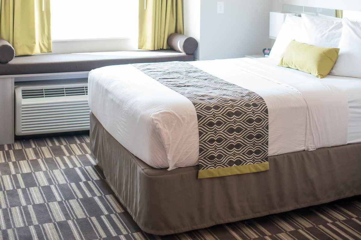 A hotel room with a bed, a window with curtains, a radiator, and a patterned carpet.
