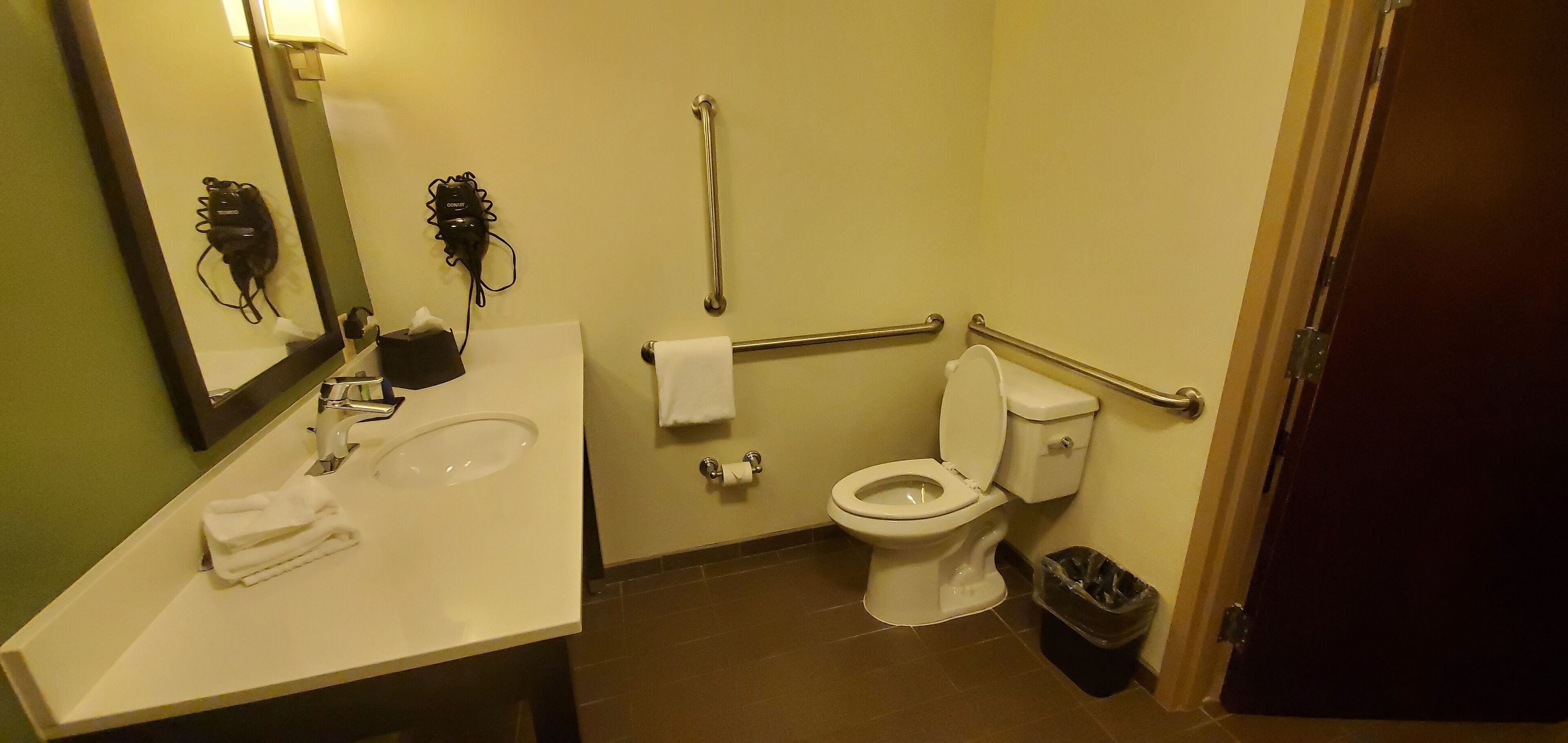 A bathroom with a sink, mirror, toilet, handrails, and a trash bin.