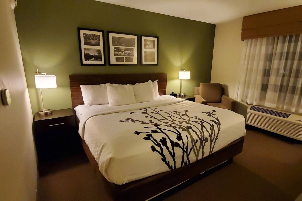 Habitación de hotel con cama, dos lámparas de noche, una silla, ventana con cortinas y equipo de aire acondicionado.