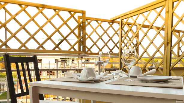 A dining table set with plates, glasses, and cutlery, with a view of an industrial area through a wooden lattice structure. - Atticoromantica (Rome)