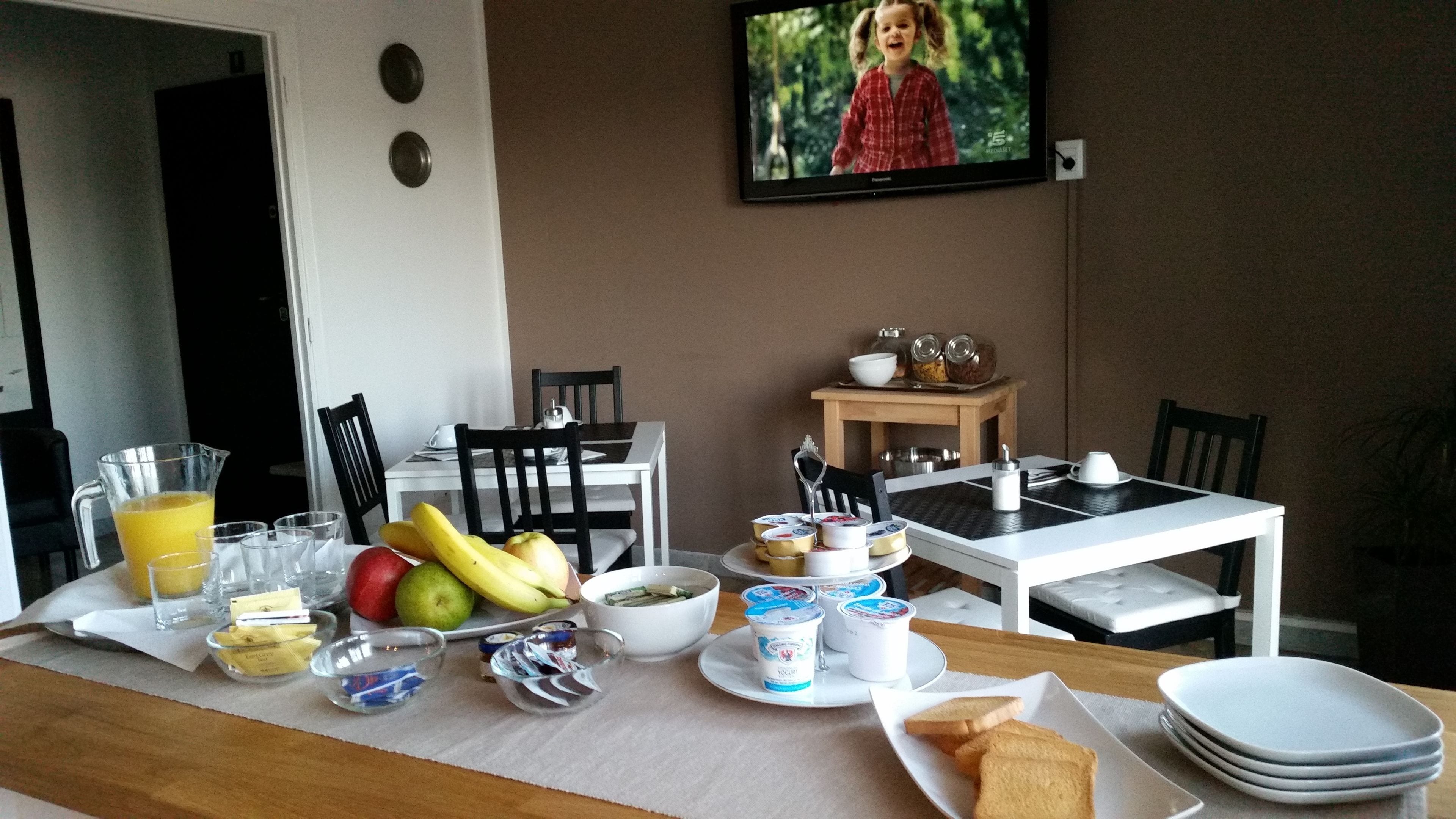 A breakfast setup with fruit, yogurt, and toast on a table, with chairs and a TV in the background.