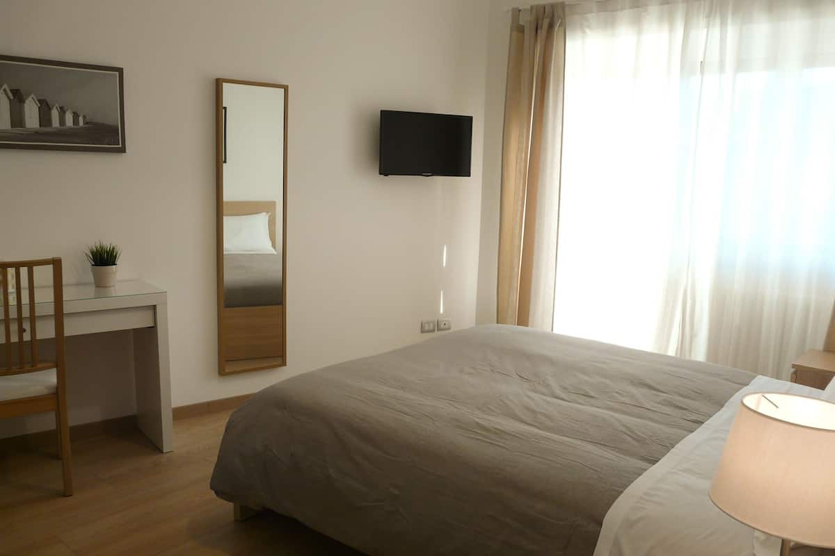 A bedroom with a bed, a desk, a chair, a television, and a window with curtains.