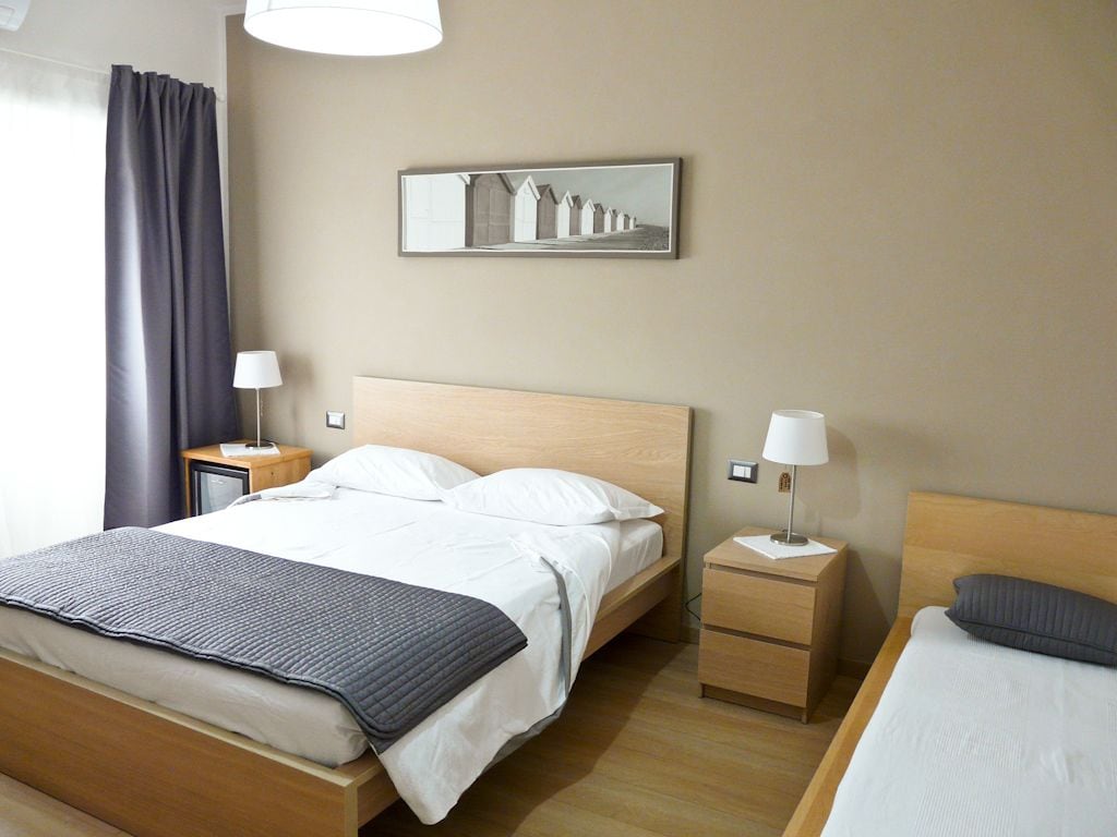 Une chambre d’hôtel avec un lit simple, une table de chevet avec une lampe, un petit tiroir et un tableau accroché au mur.
