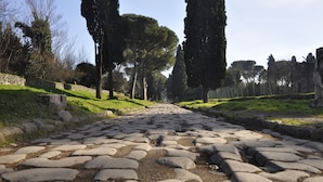 A cobblestone pathway flanked by tall trees and stone walls, leading towards a distant structure. - Atticoromantica (Rome)