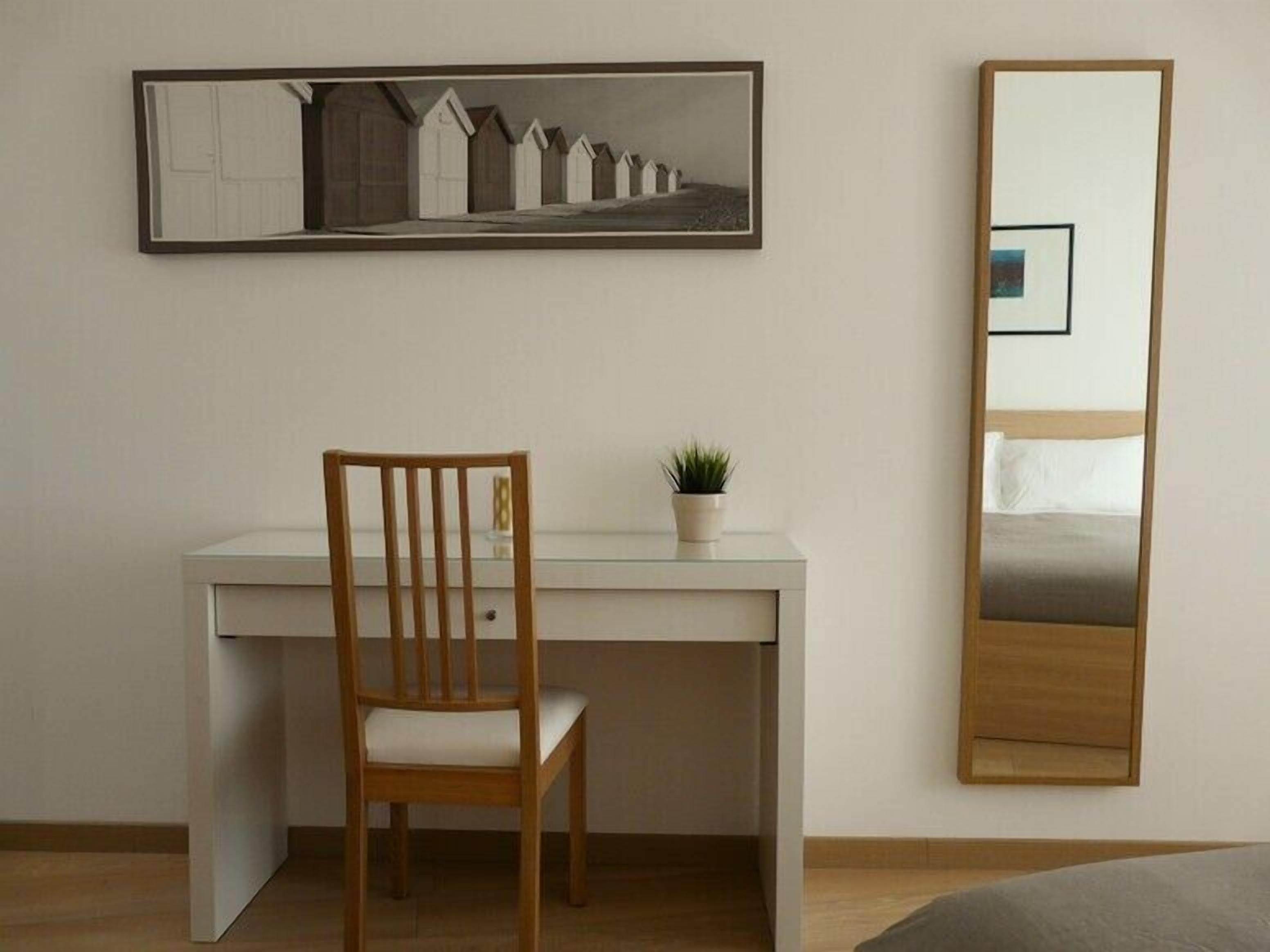 A room with a wooden chair, a desk, a potted plant, a framed picture, and a mirror.