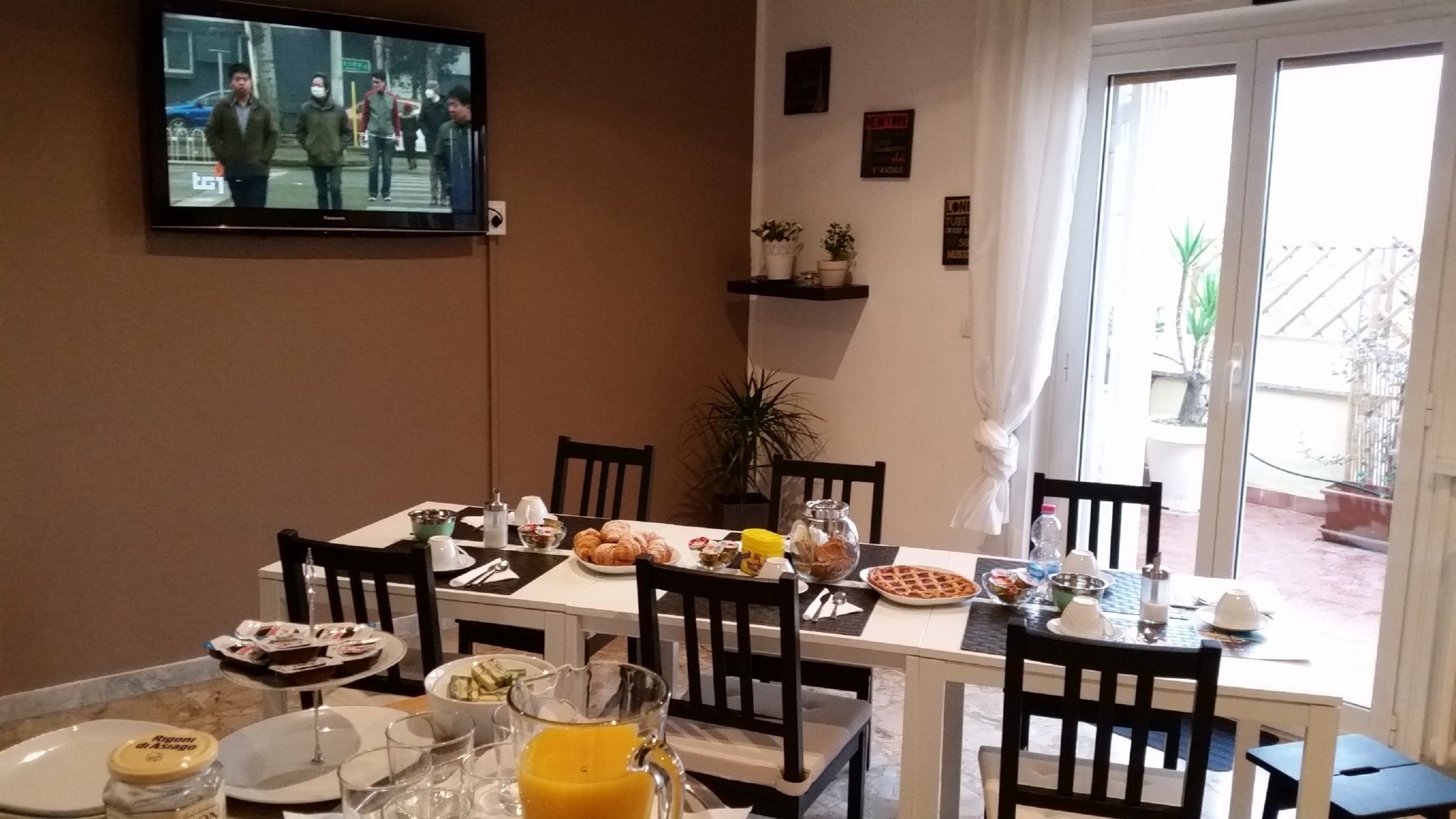 Een ontbijtruimte met een gedekte tafel, compleet met croissants, fruit en een glas sinaasappelsap. Een televisie hangt aan de muur en toont een nieuwsuitzending.