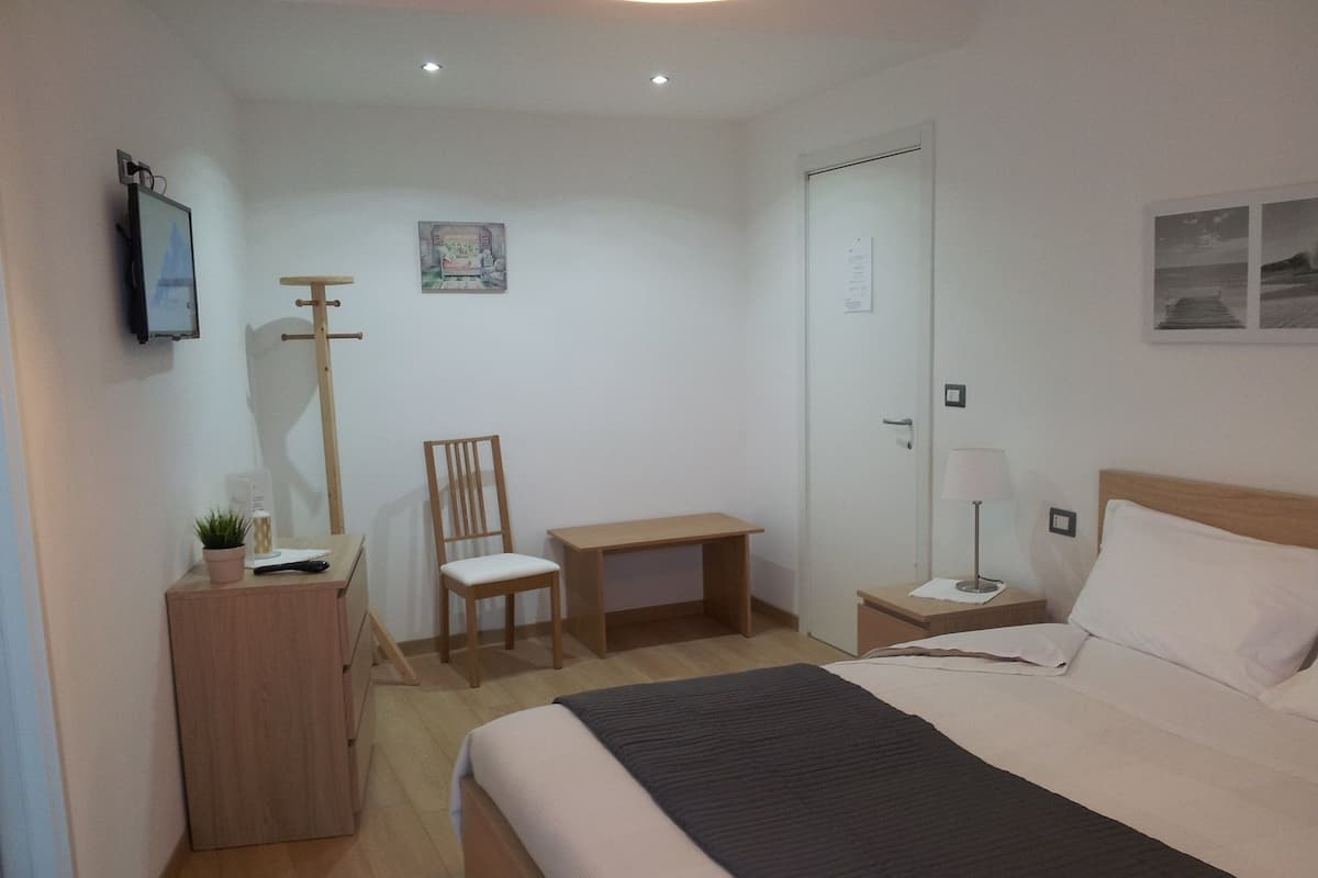 A bedroom with a bed, a desk, a chair, a bedside table with a lamp, and a wall-mounted TV.