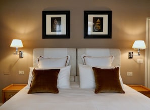 Quarto de hotel com duas camas, roupa de cama branca, almofadas castanhas e quadros nas paredes.