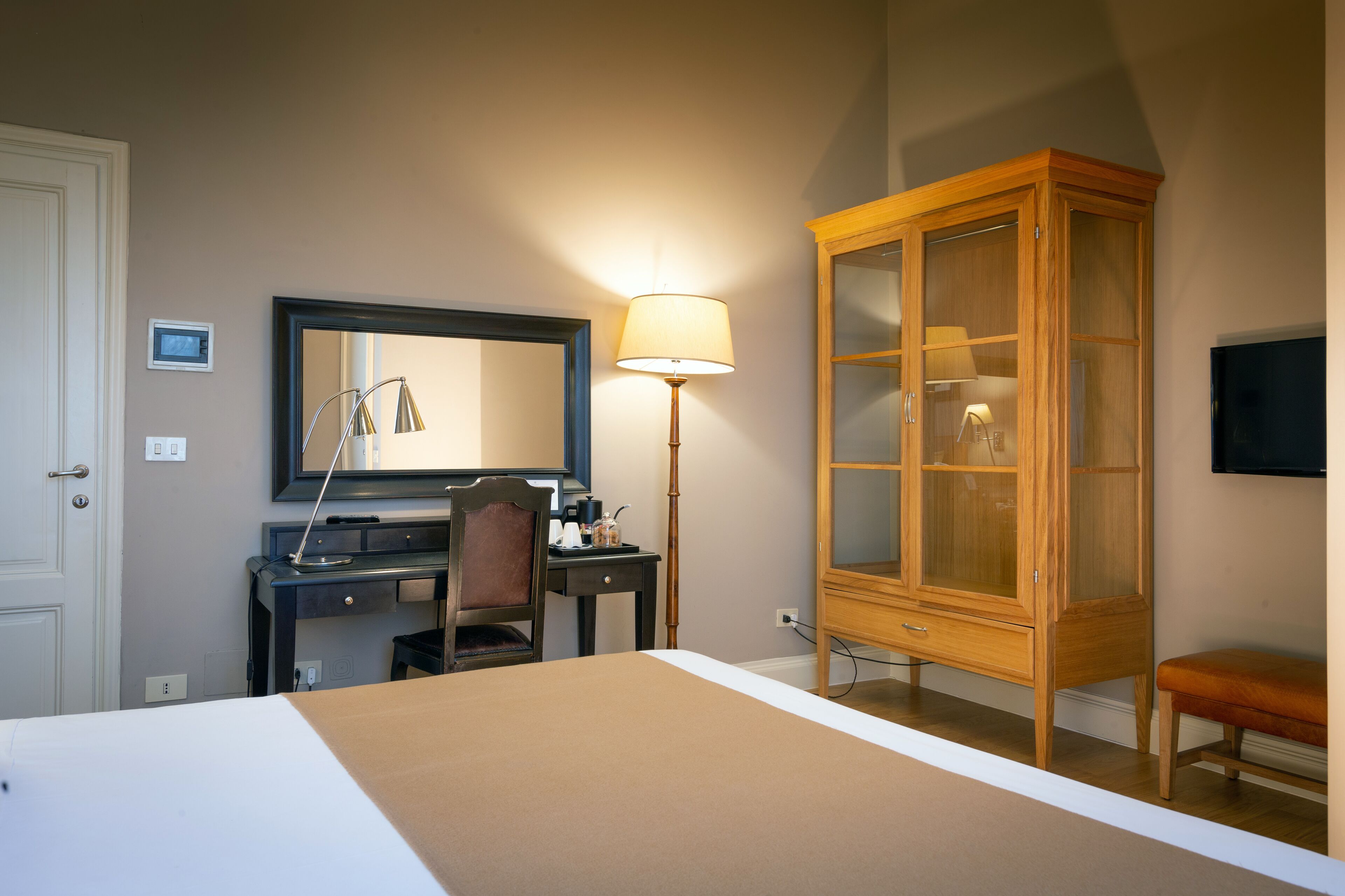 Une chambre d’hôtel avec un lit, un bureau et une chaise, une armoire en bois, une lampe et un téléviseur fixé au mur.