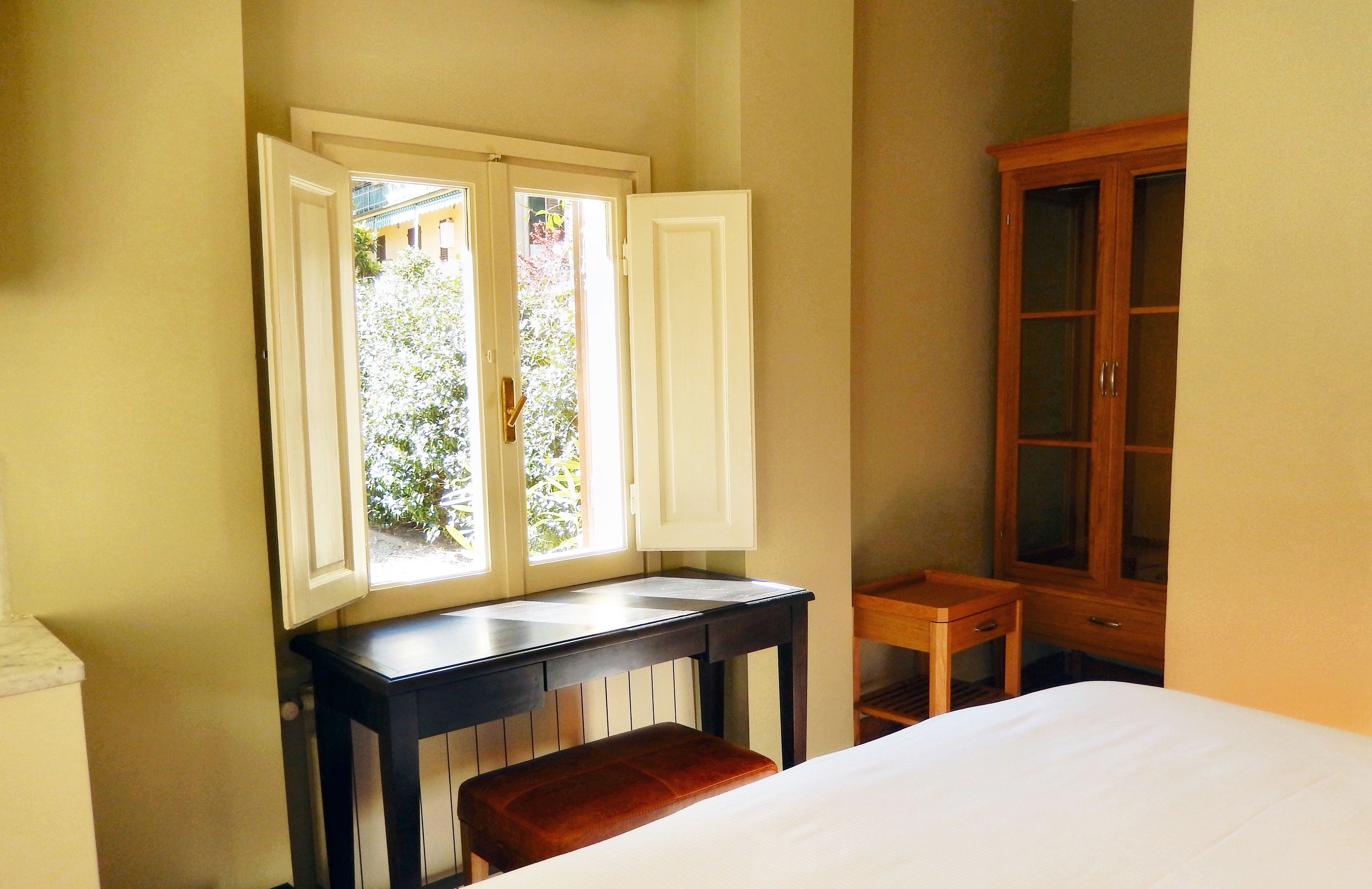 A bedroom with a bed, a wooden cabinet, a small table, and a window with open shutters.