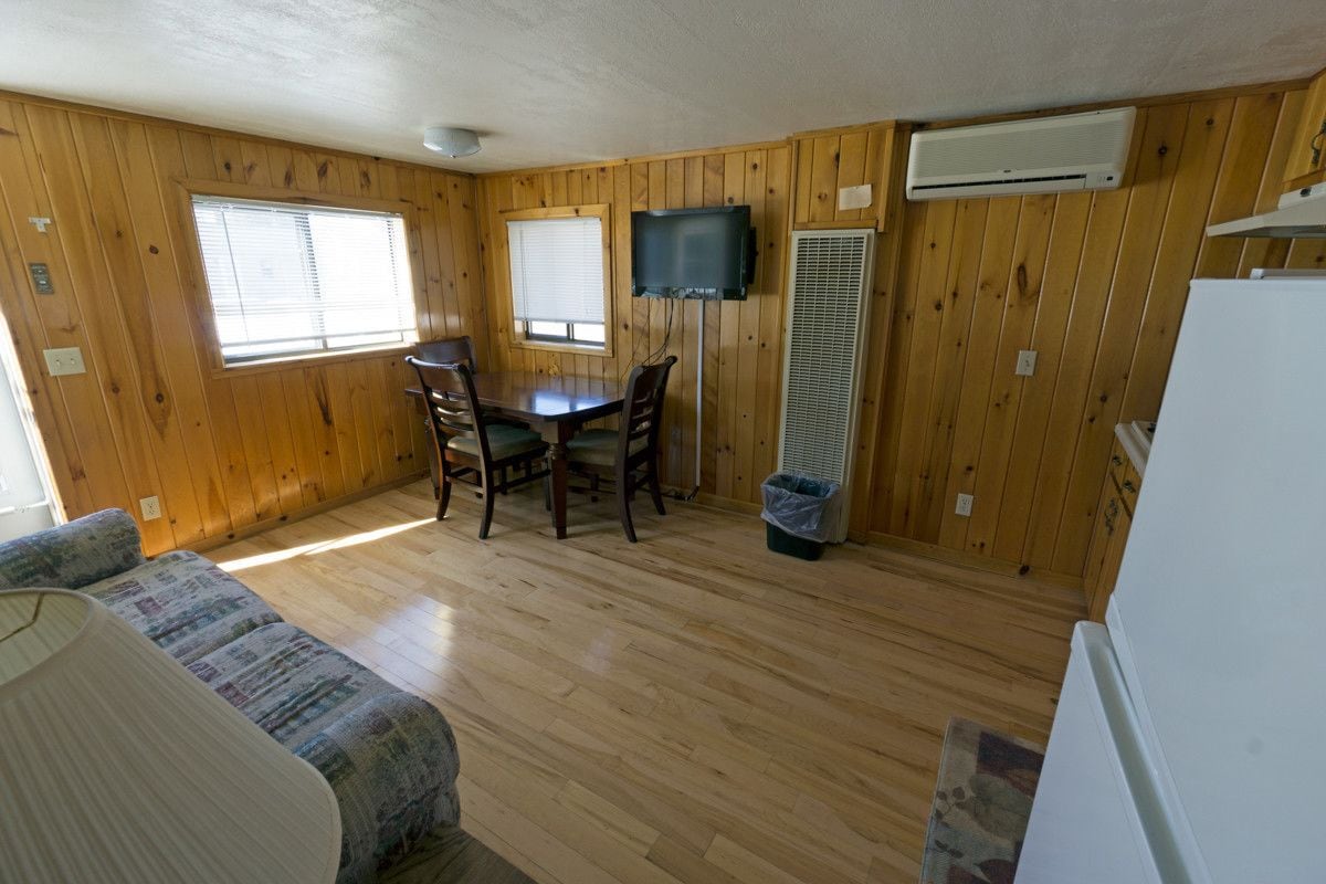 A compact room with wooden walls and floors, a dining table with chairs, a television mounted on the wall, and a white refrigerator.