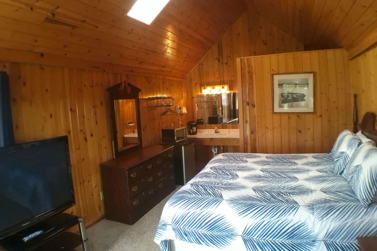 Une chambre à coucher située dans une cabane en bois, comprenant un lit, une commode, une télévision et une salle de bain avec un lavabo et un miroir.