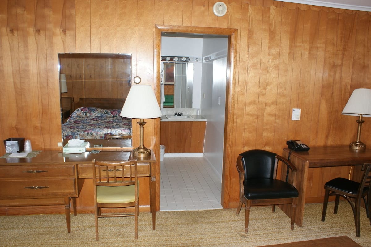 A compact hotel room with wooden paneling, a kitchenette, a bed with floral bedding, a desk with a lamp, and two chairs.
