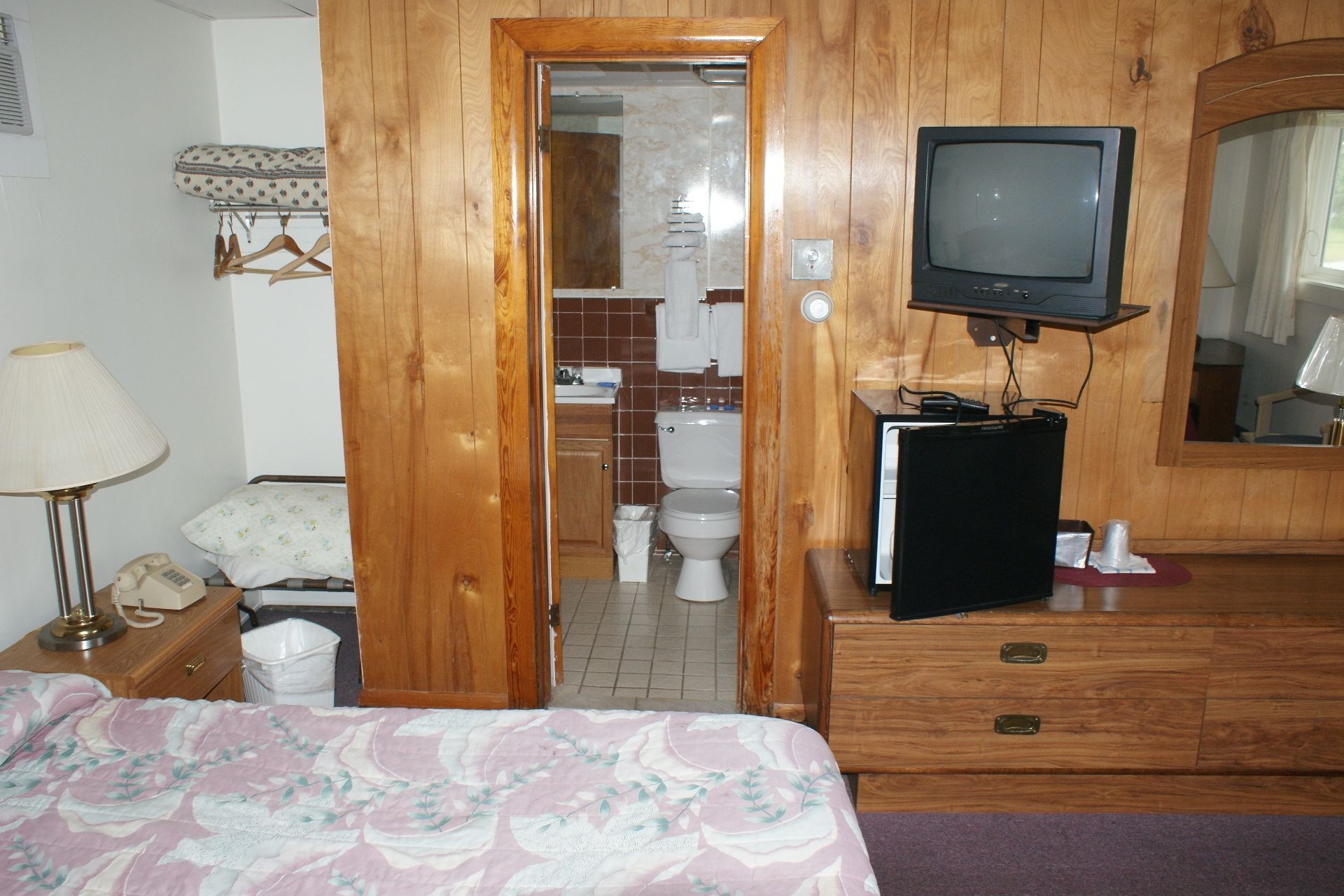 Habitación de hotel con puerta de madera, cama con colcha estampada, mesita de noche con lámpara, cómoda con espejo y televisor sobre un mueble.