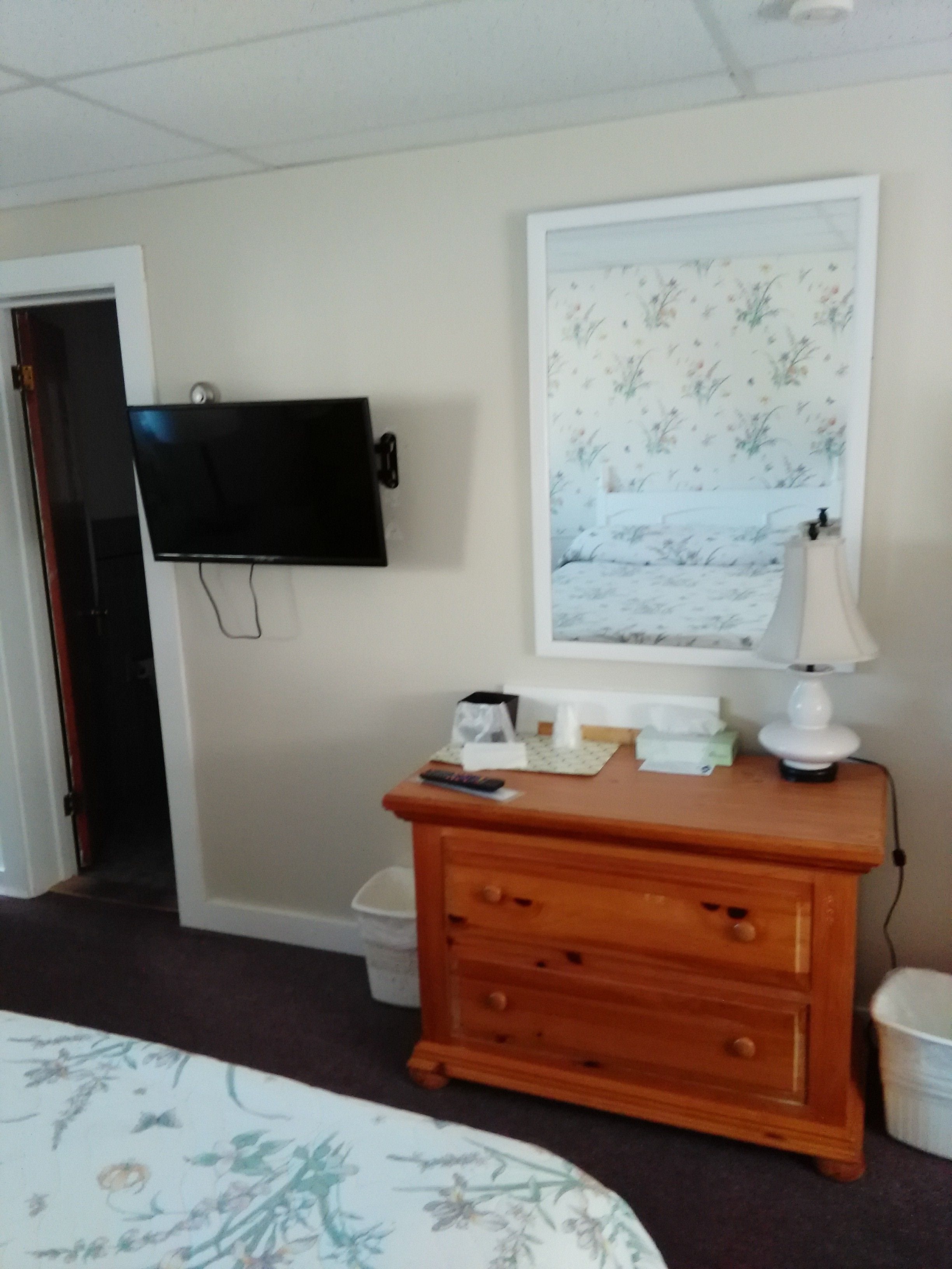 A hotel room with a bed, a wooden dresser, a television mounted on the wall, a framed floral picture, and a bedside lamp.