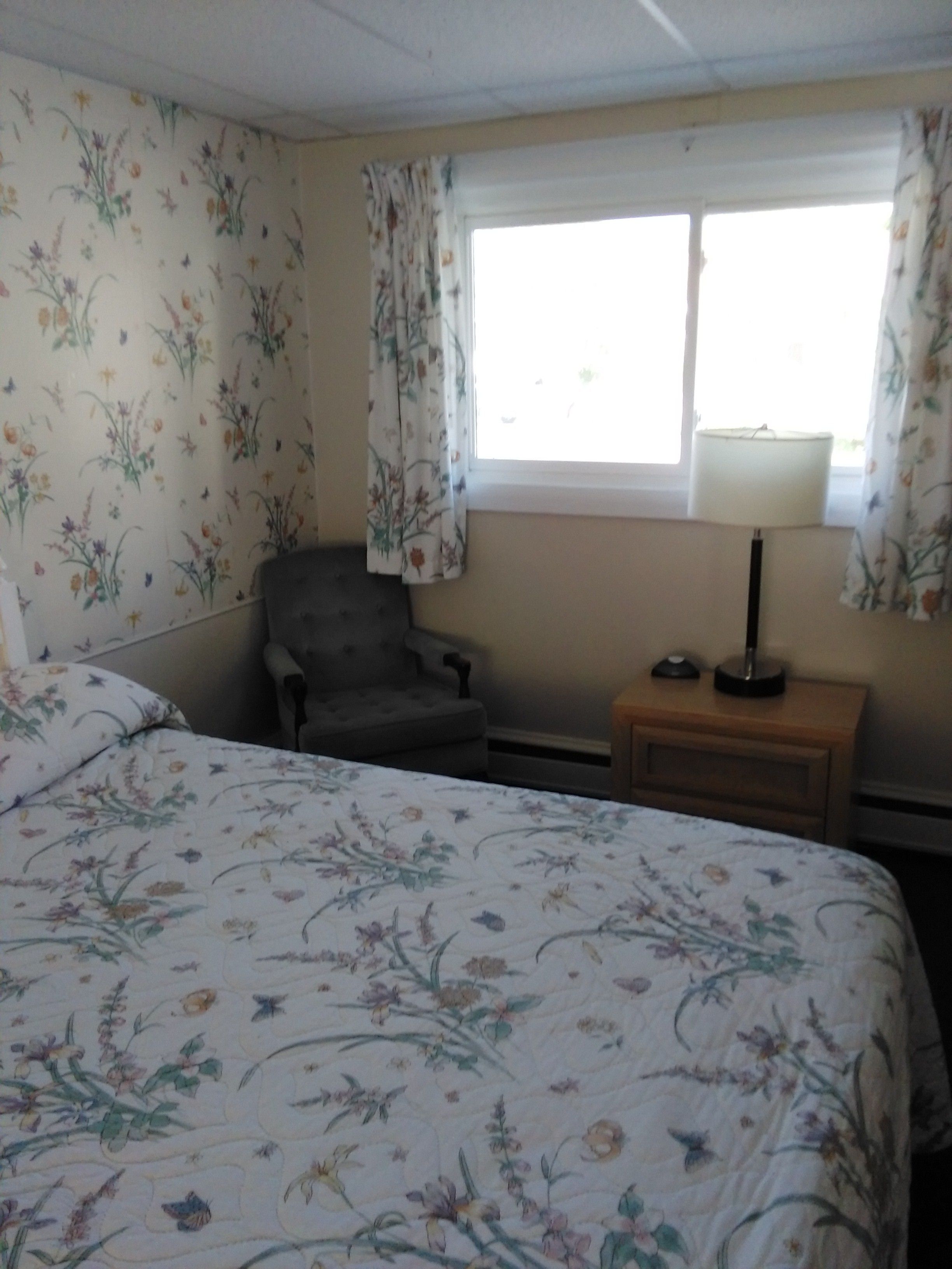 Un dormitorio con papel tapiz floral, una cama con ropa de cama estampada, un sillón gris, una mesita de noche con una lámpara y una ventana con cortinas.