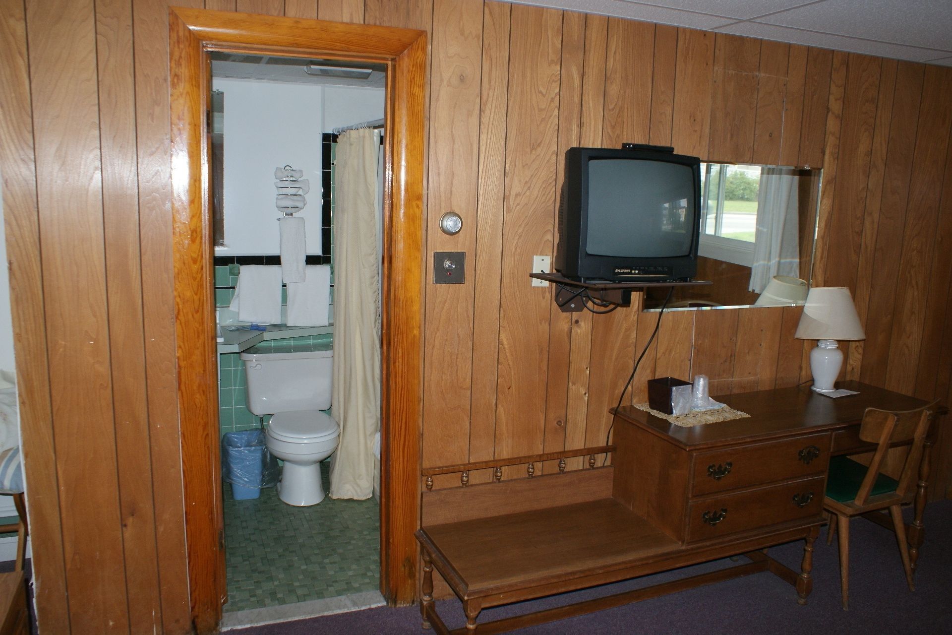 Une pièce aux murs lambrés, avec un téléviseur posé sur une étagère, un bureau avec une lampe et une chaise.