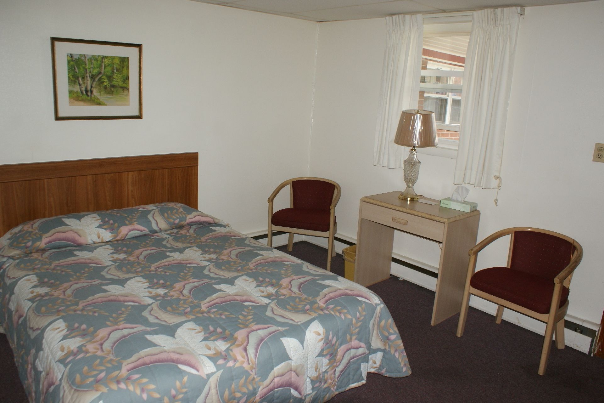 A bedroom with a bed, two chairs, a desk, a lamp, and a painting on the wall.