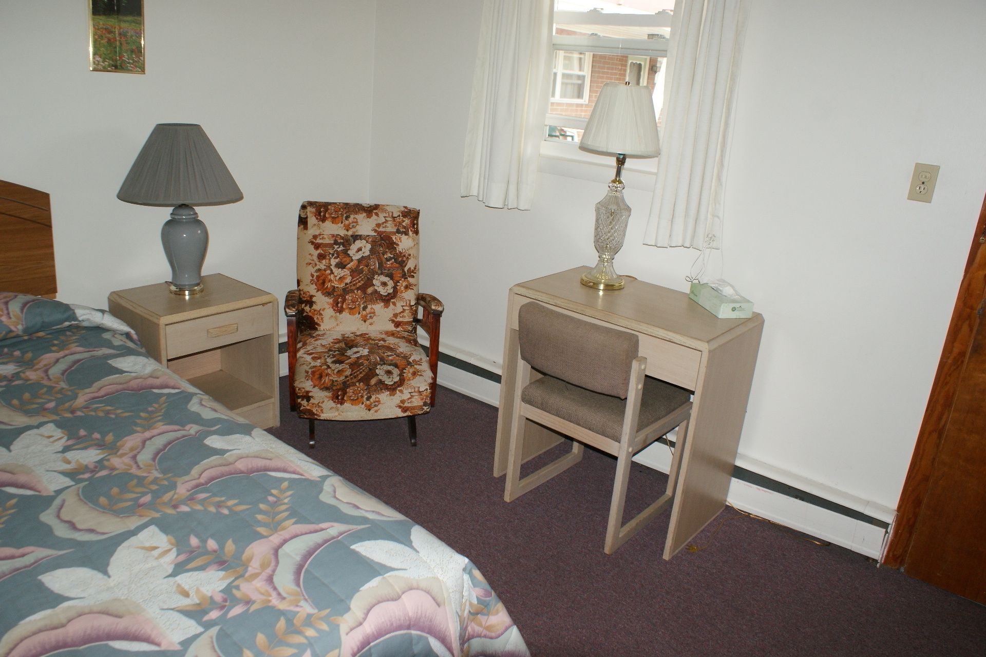 Une chambre à coucher avec un lit, un fauteuil à motifs floraux, un bureau avec une lampe et une fenêtre avec des rideaux.