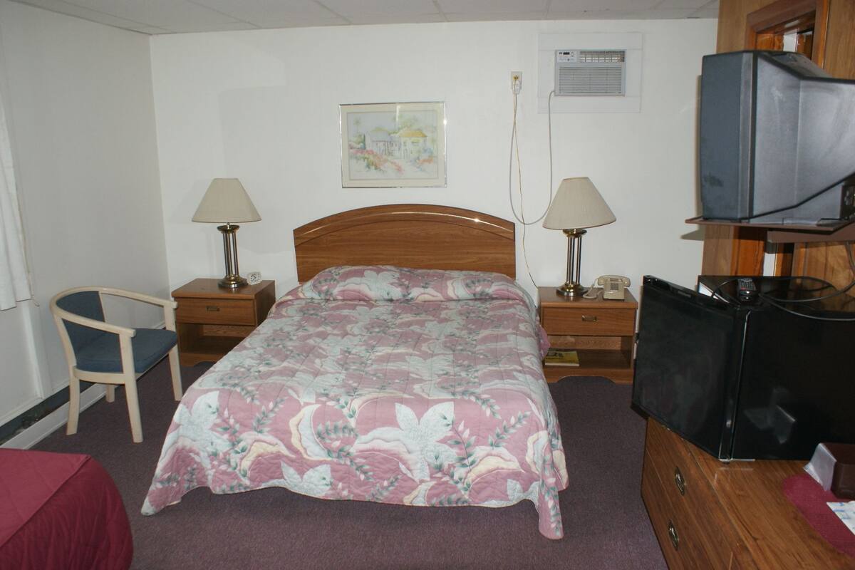 A hotel room with a bed, two bedside tables with lamps, a chair, a television, and a wall-mounted air conditioner.