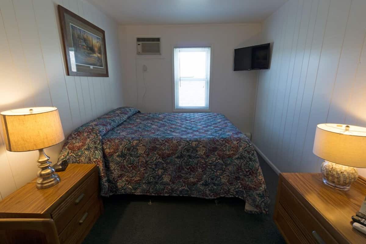 Une chambre à coucher avec un lit, deux tables de chevet avec des lampes, une fenêtre, un téléviseur fixé au mur et un tableau encadré.