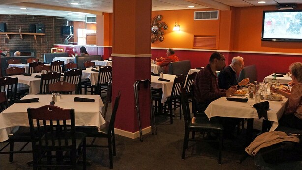 Un restaurante con varias mesas y sillas, una pantalla de televisión en la pared, y un muro de ladrillo al fondo.