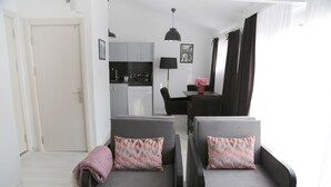 A modern living room with two grey sofas, a pink throw blanket, a dining table with chairs, a kitchen area, and a large window with curtains. - Bayer Hotel (Istanbul)