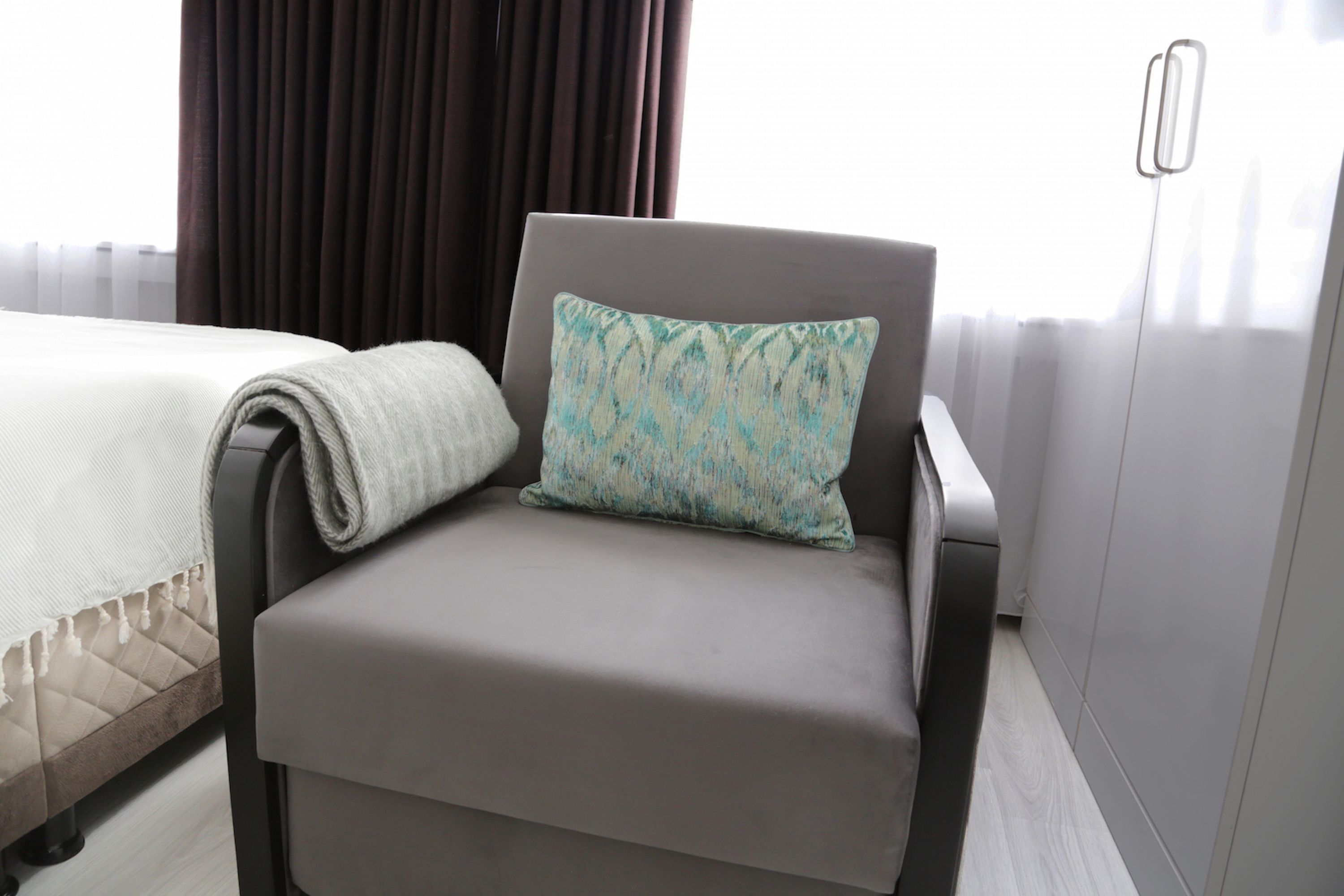 Una camera d'albergo moderna con una poltrona rivestita in grigio, un cuscino decorato e un letto rifatto.