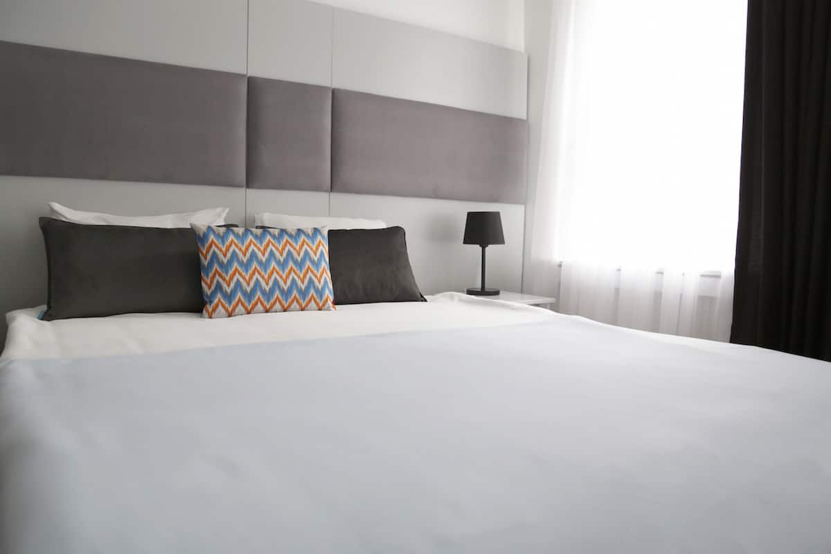 A neatly made bed with white linens and three pillows, a bedside lamp, and a window with curtains.