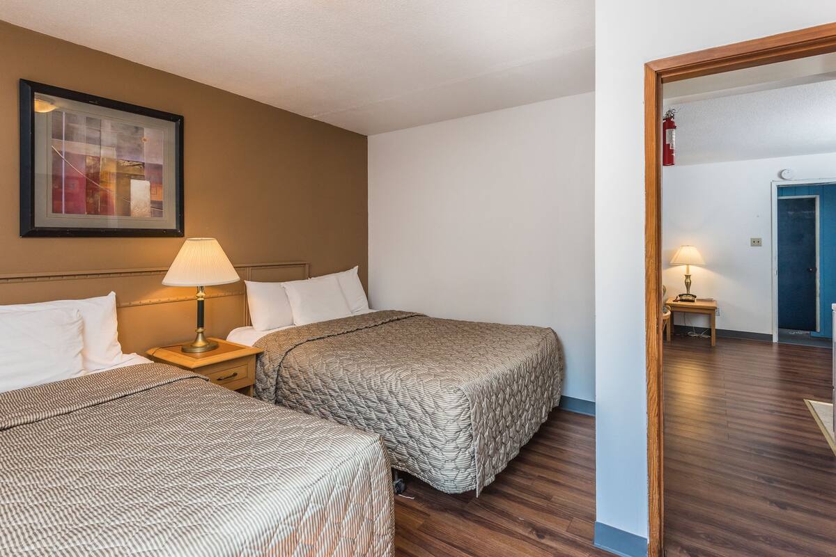 Habitación de hotel con dos camas, una mesita de noche con lámpara, un cuadro enmarcado en la pared y una puerta que da a otra habitación.