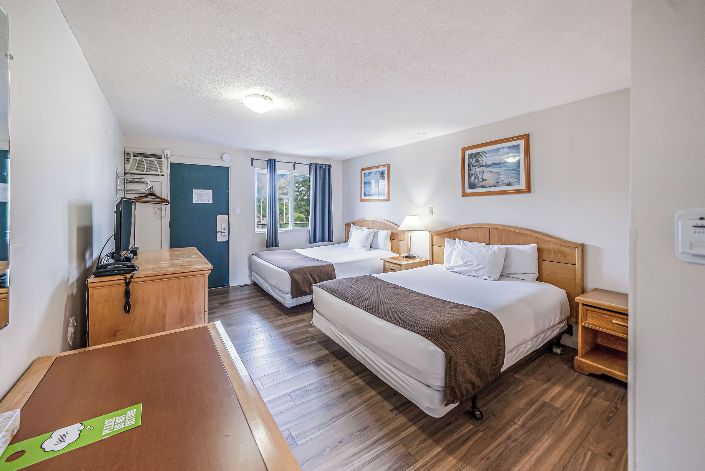 Quarto de hotel com duas camas, uma escrivaninha de madeira, mesa de cabeceira, luminária e janela com cortinas.