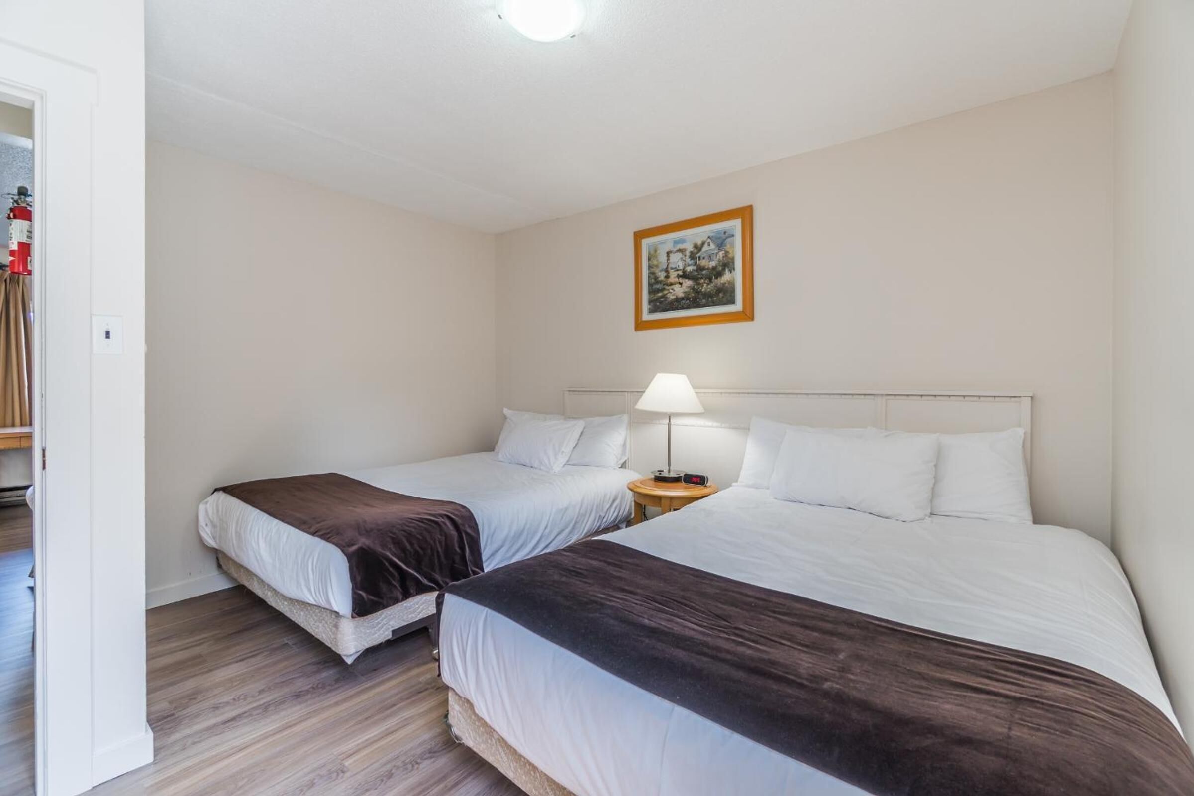 A hotel room with two beds, a bedside table with a lamp, and a framed picture on the wall.