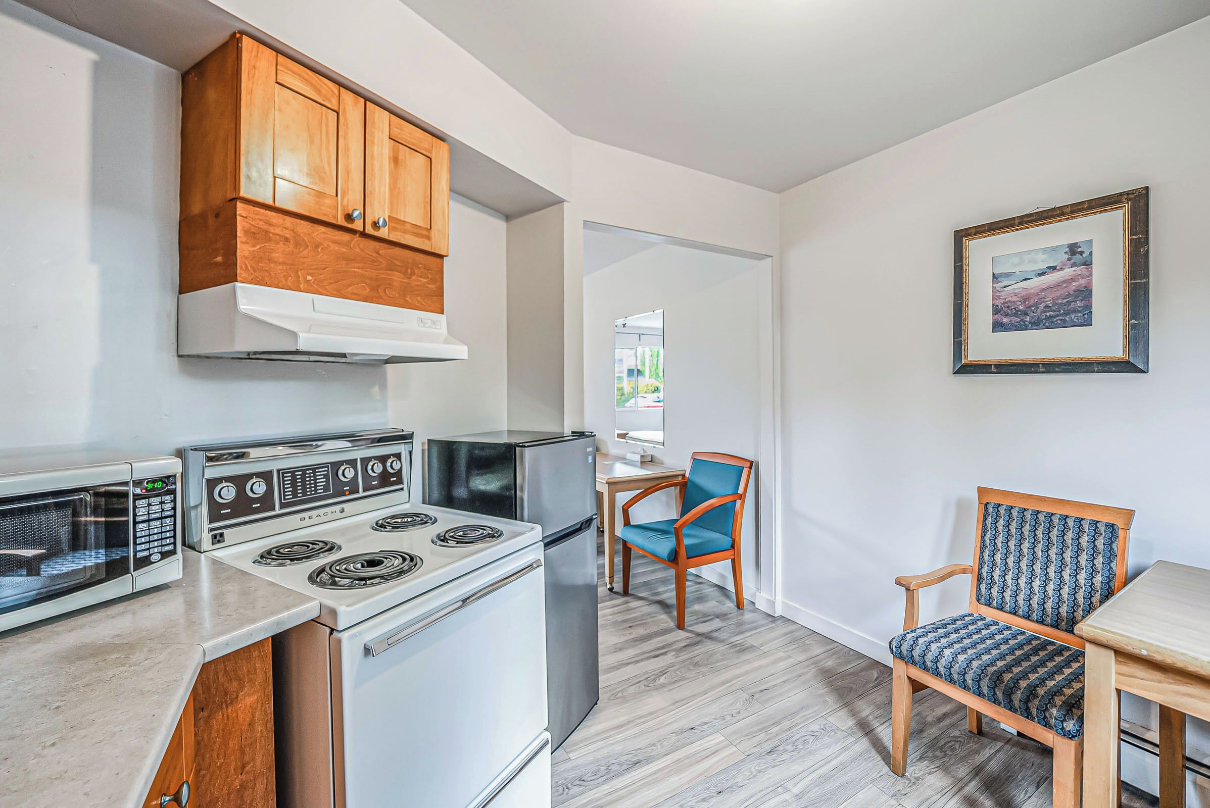 A kitchen with wooden cabinets, a stove, a refrigerator, and a microwave. There is a chair and a table in the dining area.