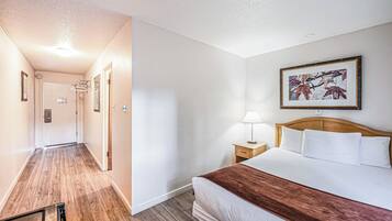 A hotel room with a wooden floor, a bed with white bedding and a brown throw, a nightstand with a lamp, and a framed picture on the wall.