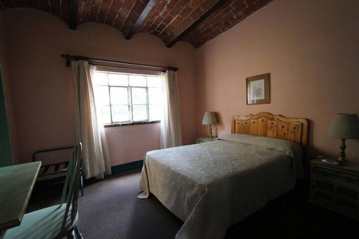 A bedroom with a single bed, a chair, a nightstand, a lamp, and a window with curtains.