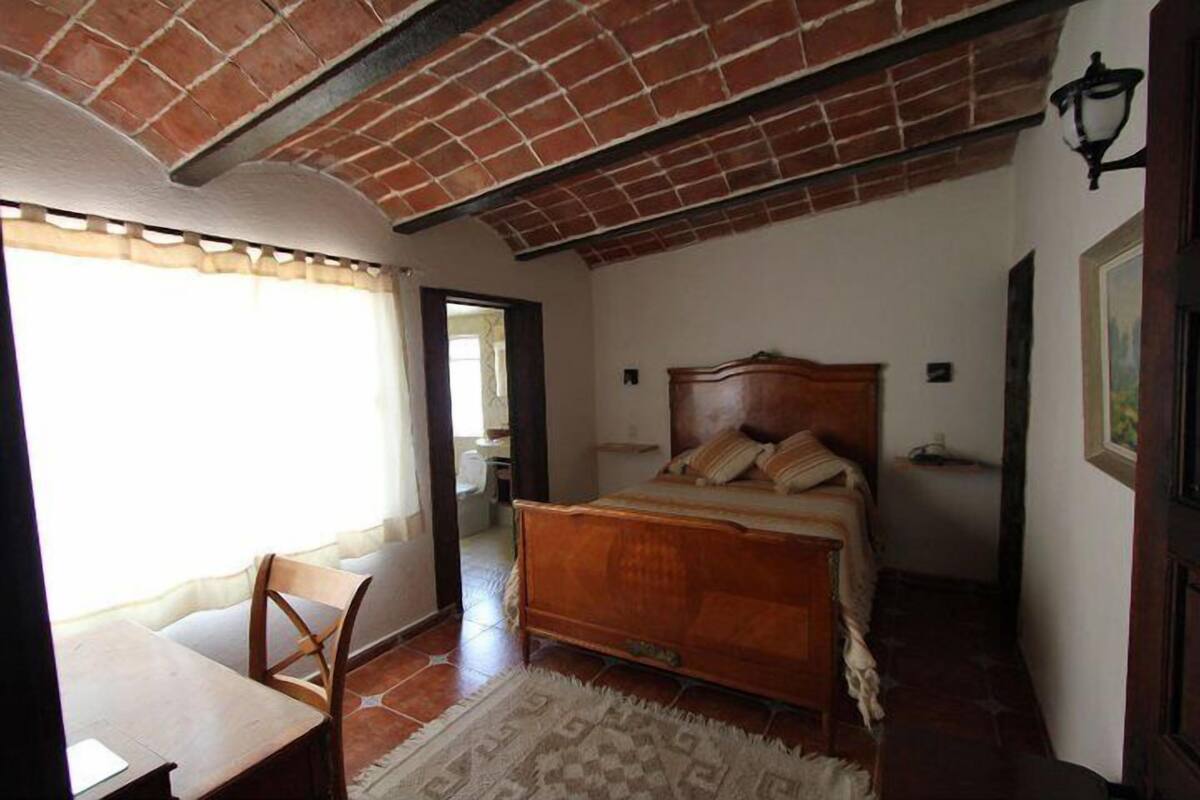 A bedroom with a wooden bed, a chair, a small table, and a patterned rug.