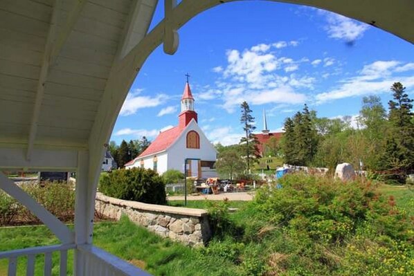 A white church with a red roof and a steeple, surrounded by greenery and trees. - Gite au Vieux Pommier (Tadoussac)