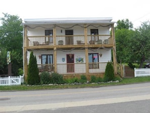 A two-story building with a balcony on the second floor, a white picket fence, and a road in front. - Gite au Vieux Pommier (Tadoussac)